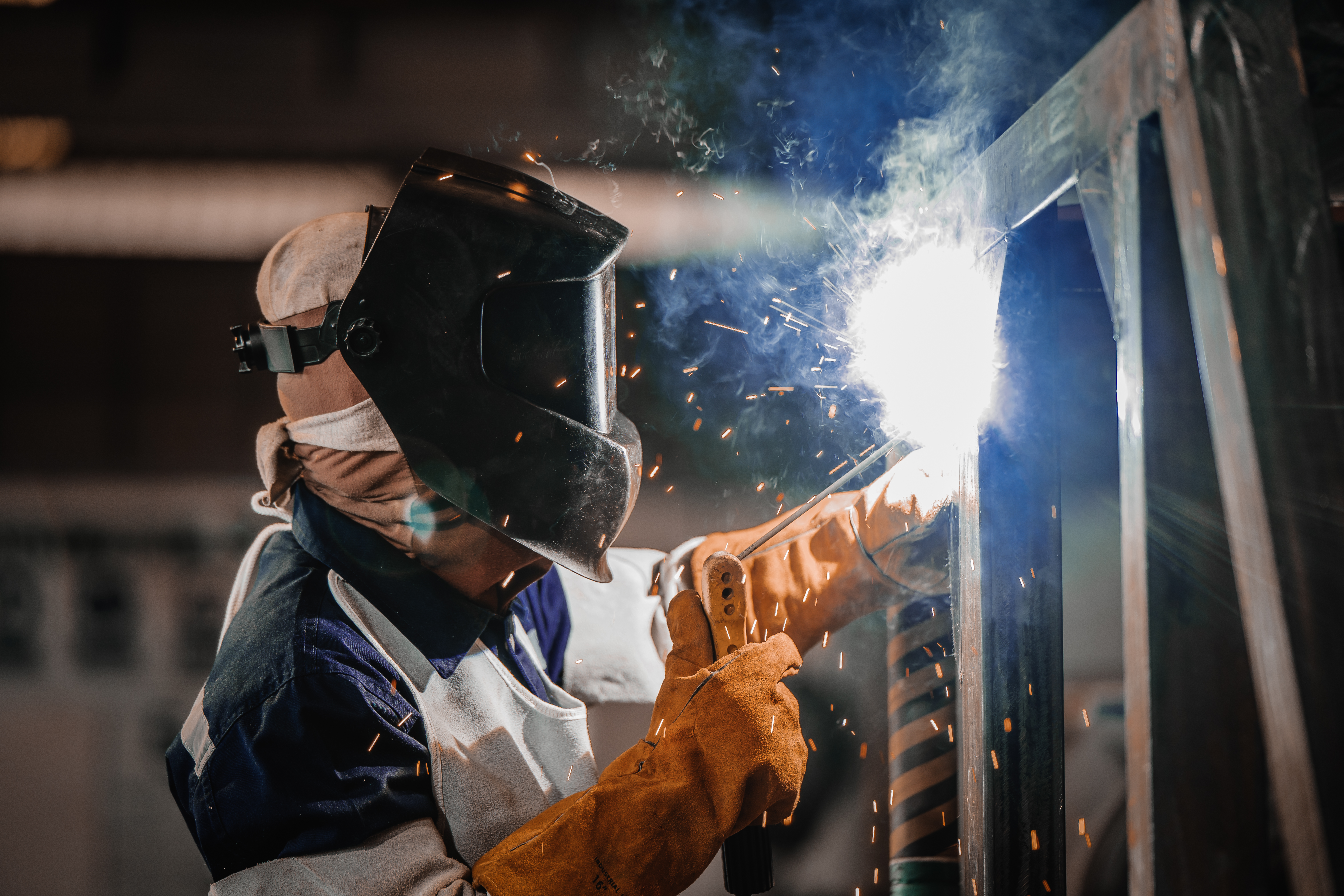 Industrial worker welding steel structure sparks protective mask leather gloves. Manual arc welding metal frame factory production line heavy industry metalwork fabrication process steel.